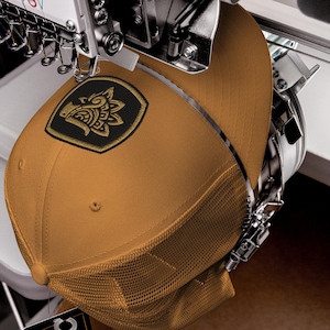Puede incluir: Gorra de camionero de color mostaza con un parche bordado en negro y dorado con un diseño estilizado. La gorra tiene la parte trasera de malla y está siendo trabajada por una máquina de bordar.