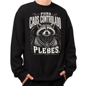 May include: A black sweatshirt featuring a graphic print of a raccoon wearing a bandana and a chain with a paw print pendant. The text PURO CAOS CONTROLADO PLEBES is printed in white above and below the raccoon, surrounded by decorative flourishes.