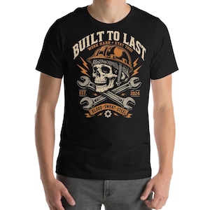 Könnte beinhalten: Ein schwarzes T-Shirt mit einem Grafikdruck eines Schädels mit Schutzhelm, der über zwei gekreuzten Schraubenschlüsseln abgebildet ist. Der Text lautet BUILT TO LAST, WORK HARD STAY TRUE, EST. 2024 und BLOOD SWEAT STEEL in Orange und Creme.
