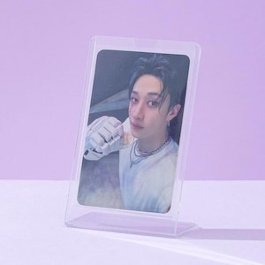 Puede incluir: Un soporte fotográfico de plástico transparente que muestra una fotografía de una persona con cabello oscuro, que lleva un suéter blanco y múltiples collares plateados. El soporte fotográfico está sobre una superficie blanca sobre un fondo morado.