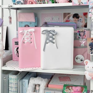 Pode incluir: Um caderno rosa e branco com um design de atacadores estilo espartilho. Os cadernos são exibidos numa prateleira branca com vários outros itens, incluindo peluches, cartões de fotos e caixas decorativas. A prateleira faz parte de uma coleção.