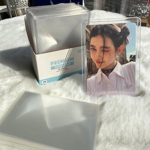 May include: A stack of clear plastic photocard top loaders, 65 x 85 mm, with a photocard inside one of the top loaders. The top loader has a clear plastic front and back and a white border. The photocard shows a young woman with dark hair and a white shirt. The photocard is in a clear plastic top loader. The top loader is labeled "Premium Toploader 65 x 85 mm Photocard".