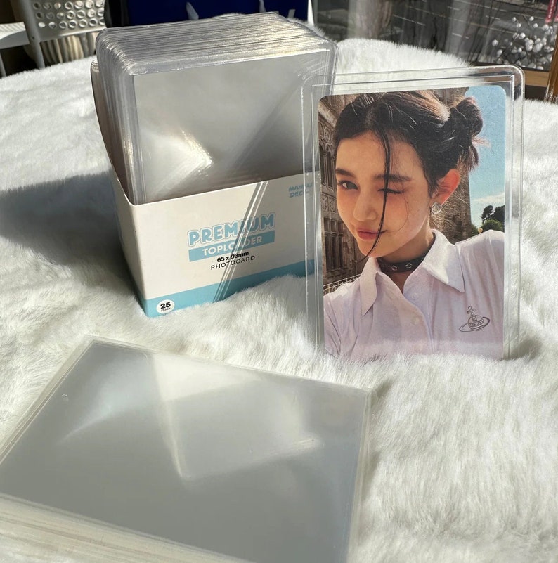 May include: A stack of clear plastic photocard top loaders, 65 x 85 mm, with a photocard inside one of the top loaders. The top loader has a clear plastic front and back and a white border. The photocard shows a young woman with dark hair and a white shirt. The photocard is in a clear plastic top loader. The top loader is labeled "Premium Toploader 65 x 85 mm Photocard".