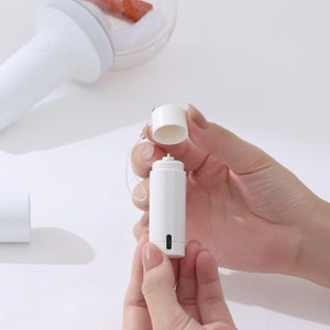 May include: A white cylindrical container with a white lid and a small black charging port. The container has a white string attached to the lid.