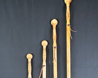 May include: Four wooden walking sticks of varying heights, each featuring a rounded top and a slender shaft. The sticks are crafted from light-colored wood, with intricate carvings on some. Leather cords are attached near the top of each stick.