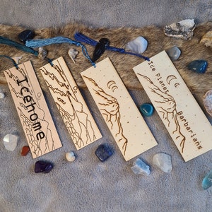 May include: Four wooden bookmarks with different designs and text. The bookmarks are on a furry surface with various small stones and shells. The text on the bookmarks reads "Ice Planet Barbarians", "Oo. Teehome", "Oo." and "Oo."