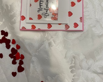 Può includere: Un biglietto di San Valentino rosa e bianco con una finestra trasparente che mostra un design a forma di cuore scintillante. Il biglietto ha il testo "Happy Valentine's" scritto su di esso.