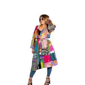 K&ouml;nnte beinhalten: Ein langer, bunter Patchwork-Mantel mit einem Bindeg&uuml;rtel. Der Mantel besteht aus verschiedenen Stoffen und Mustern, darunter Blumen, geometrische Muster und Paisley. Das Model tr&auml;gt den Mantel mit Jeans und schwarzen Schuhen.