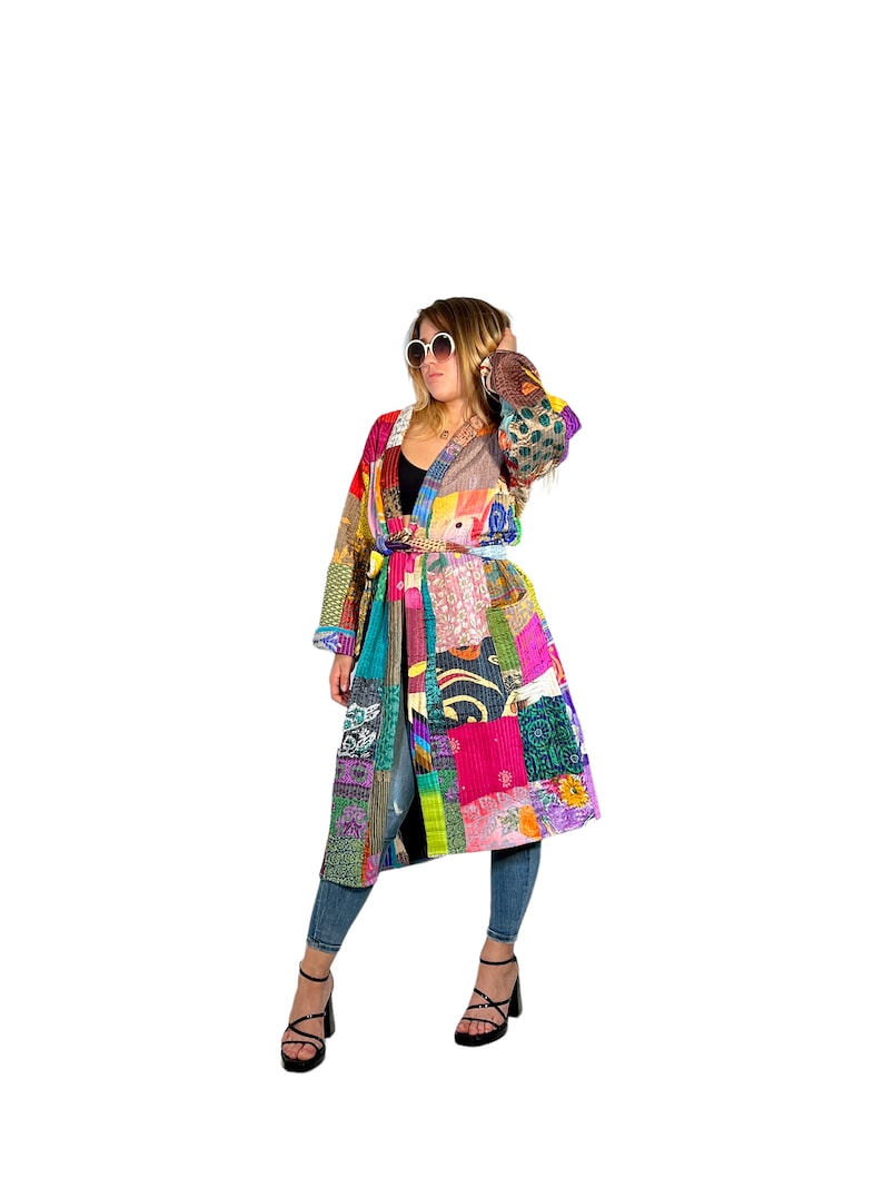 K&ouml;nnte beinhalten: Ein langer, bunter Patchwork-Mantel mit einem Bindeg&uuml;rtel. Der Mantel besteht aus verschiedenen Stoffen und Mustern, darunter Blumen, geometrische Muster und Paisley. Das Model tr&auml;gt den Mantel mit Jeans und schwarzen Schuhen.