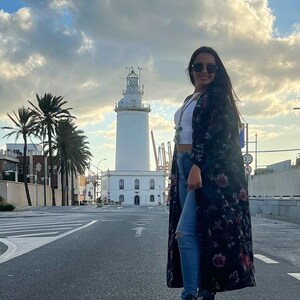 Puede incluir: Una mujer con un abrigo largo negro con estampado floral, top corto blanco y vaqueros azules est&aacute; de pie en una calle. Lleva gafas de sol y zapatillas altas negras. Un faro blanco se encuentra en el fondo bajo un cielo nublado.