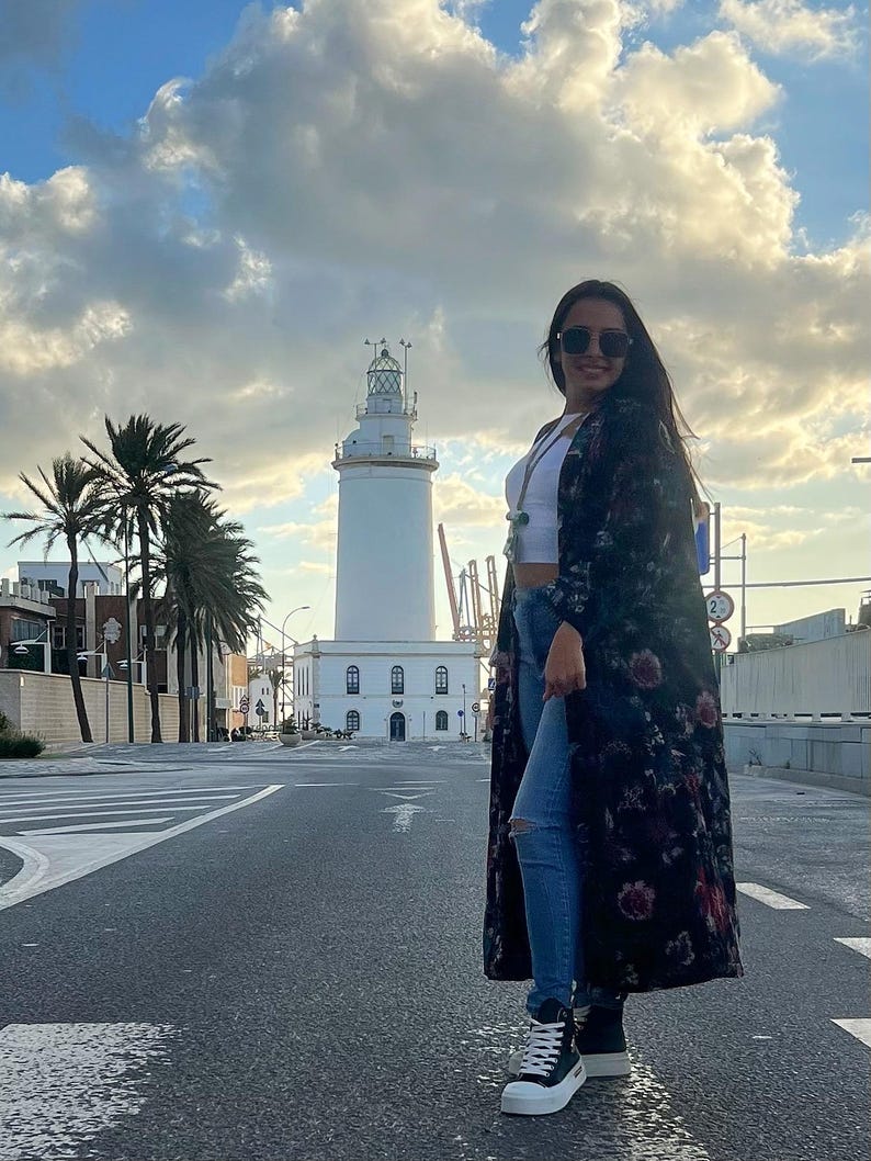 Puede incluir: Una mujer con un abrigo largo negro con estampado floral, top corto blanco y vaqueros azules est&aacute; de pie en una calle. Lleva gafas de sol y zapatillas altas negras. Un faro blanco se encuentra en el fondo bajo un cielo nublado.