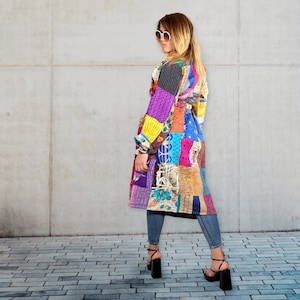 Puede incluir: Una larga chaqueta de patchwork colorida con cinturón. La chaqueta está hecha de diferentes telas y patrones en tonos de morado, azul, verde, amarillo y rojo. La modelo lleva la chaqueta con jeans y tacones negros.