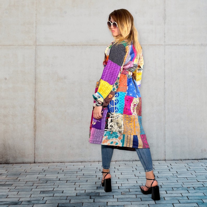 K&ouml;nnte beinhalten: Ein langer, farbenfroher Patchwork-Mantel mit G&uuml;rtel. Der Mantel besteht aus verschiedenen Stoffen und Mustern in den Farben Lila, Blau, Gr&uuml;n, Gelb und Rot. Das Model tr&auml;gt den Mantel mit Jeans und schwarzen Schuhen.
