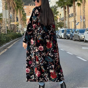 Puede incluir: Abrigo largo negro con estampado floral en rojo, rosa y verde. Se lleva sobre vaqueros azules y zapatillas de plataforma blancas y negras. El abrigo tiene una textura de terciopelo.