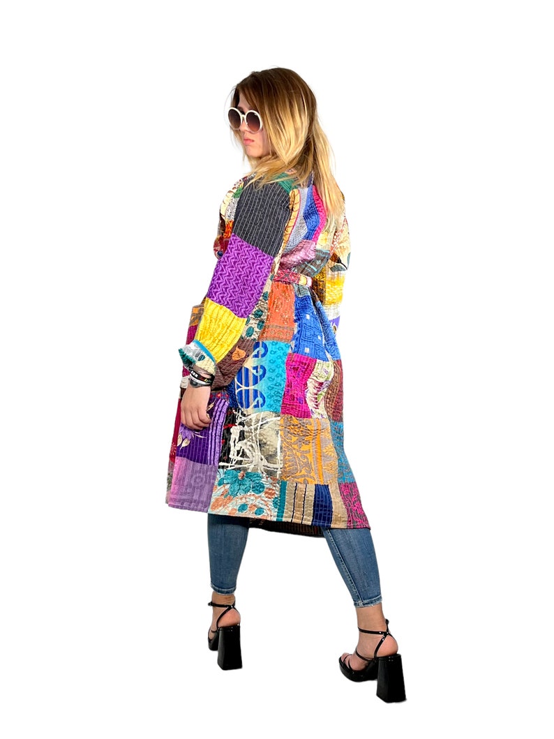 K&ouml;nnte beinhalten: Ein langer, bunter Patchwork-Mantel mit G&uuml;rtel. Der Mantel besteht aus verschiedenen Stoffen und Mustern, darunter Blumen, geometrische Muster und Paisley. Das Model tr&auml;gt den Mantel mit Jeans und schwarzen Schuhen.