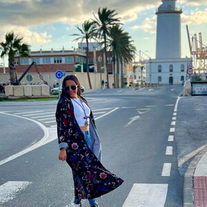 Puede incluir: Una mujer con un kimono largo con estampado floral oscuro, top blanco, vaqueros azules y zapatillas deportivas blancas y negras. El fondo muestra un faro, palmeras y un cielo nublado.