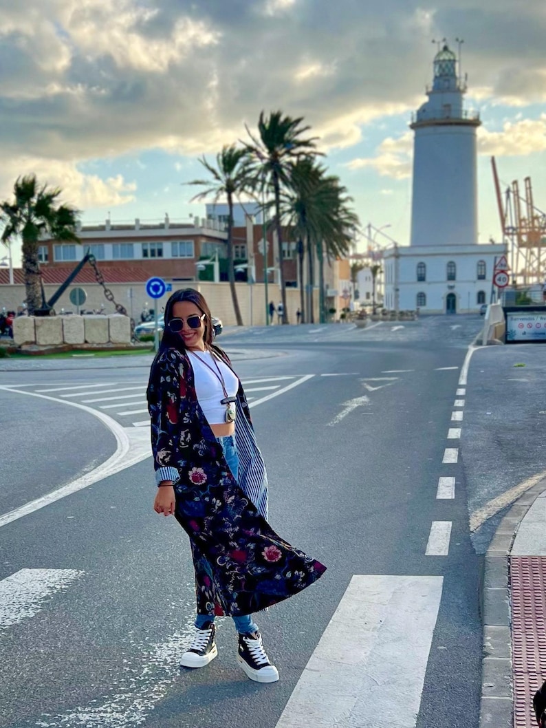 Puede incluir: Una mujer con un kimono largo con estampado floral oscuro, top blanco, vaqueros azules y zapatillas deportivas blancas y negras. El fondo muestra un faro, palmeras y un cielo nublado.