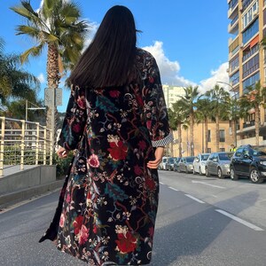 Puede incluir: Abrigo largo negro con estampado floral con flores rojas y rosas, hojas verdes y pu&ntilde;o a rayas. El abrigo se lleva al aire libre en un d&iacute;a soleado.
