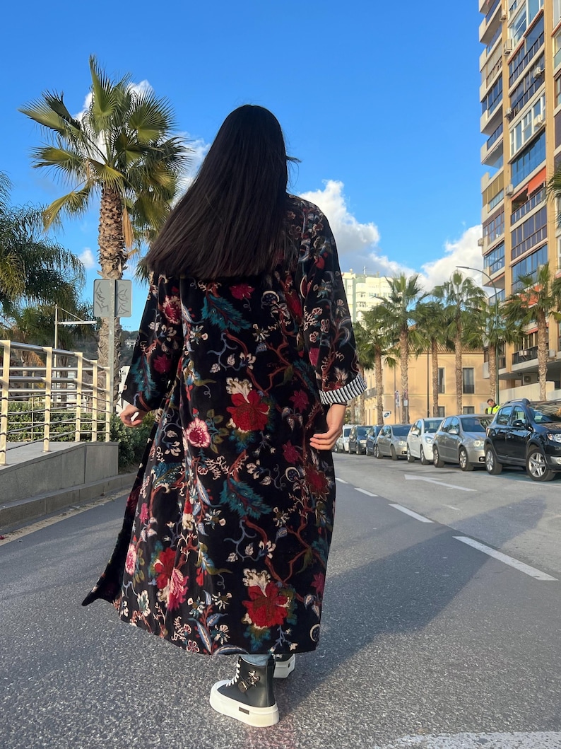 Puede incluir: Abrigo largo negro con estampado floral con flores rojas y rosas, hojas verdes y pu&ntilde;o a rayas. El abrigo se lleva al aire libre en un d&iacute;a soleado.