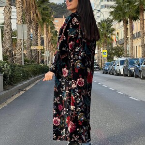 Puede incluir: Abrigo largo negro con estampado floral con detalles en rojo, rosa y verde. El abrigo se lleva con botas blancas y negras. La persona lleva gafas de sol y est&aacute; de pie en una calle bordeada de palmeras.
