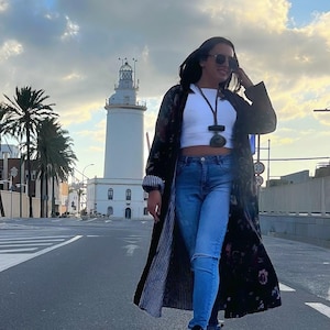 Puede incluir: Una mujer con un abrigo largo negro con estampado floral, top corto blanco y vaqueros azules. Lleva zapatillas altas negras y blancas y gafas de sol. Un faro blanco se encuentra al fondo.