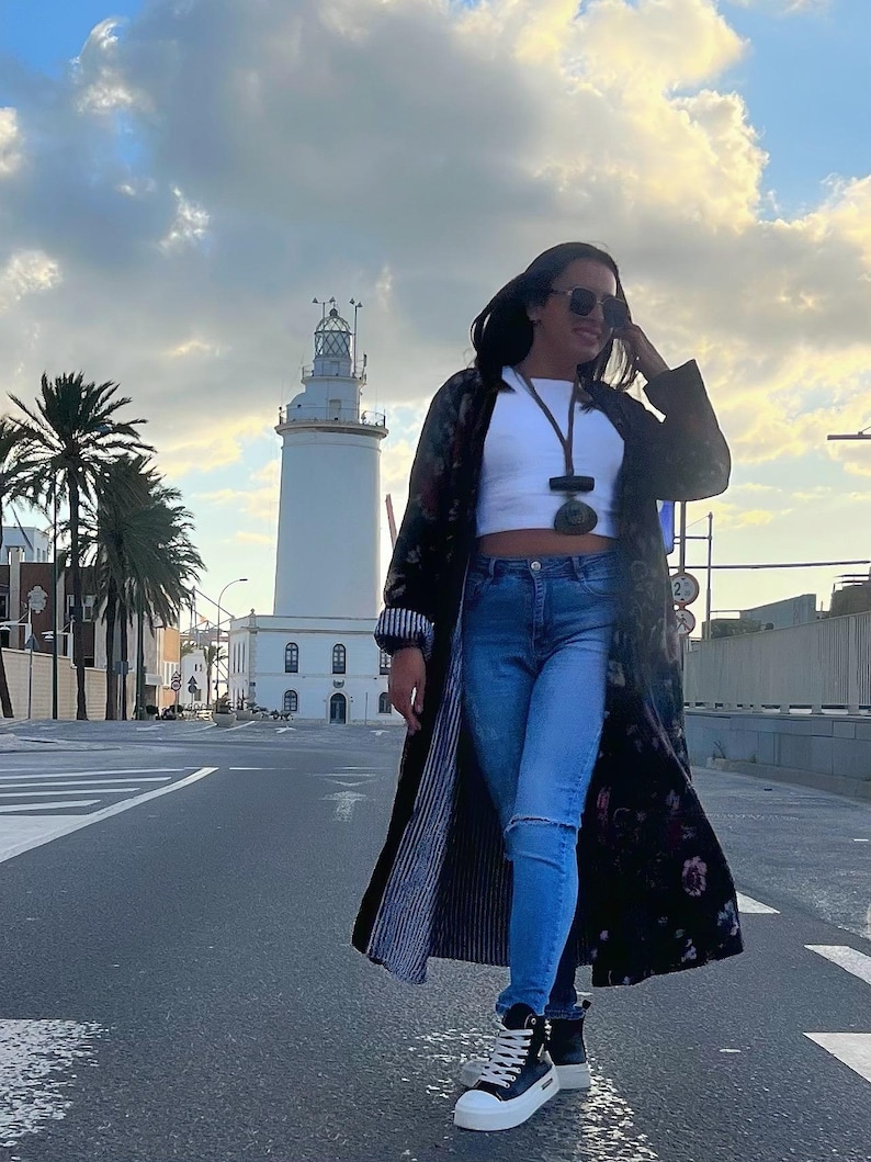 Puede incluir: Una mujer con un abrigo largo negro con estampado floral, top corto blanco y vaqueros azules. Lleva zapatillas altas negras y blancas y gafas de sol. Un faro blanco se encuentra al fondo.