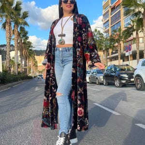 Puede incluir: Una mujer con un kimono largo negro con estampado floral, top blanco y vaqueros rotos azul claro. Tambi&eacute;n lleva zapatillas altas negras y gafas de sol. El fondo muestra palmeras y una calle de la ciudad.