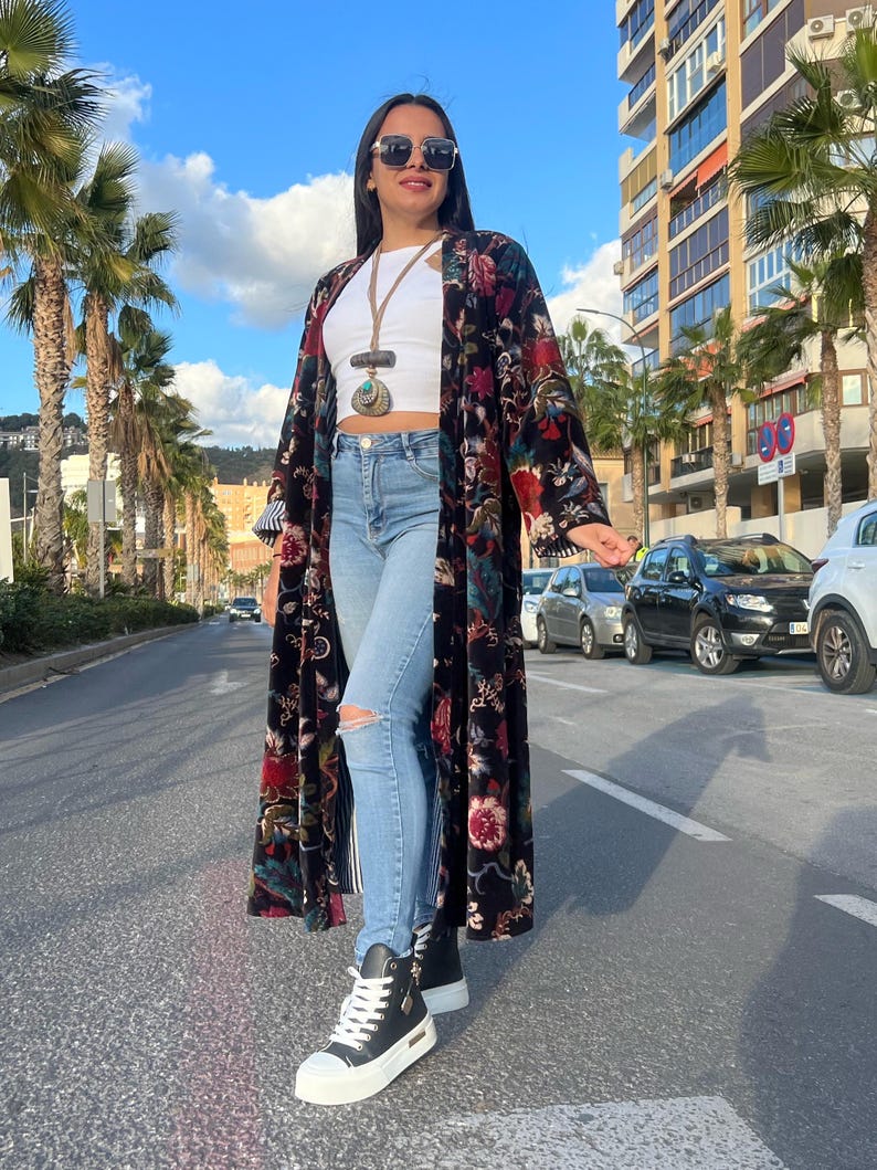 Puede incluir: Una mujer con un kimono largo negro con estampado floral, top blanco y vaqueros rotos azul claro. Tambi&eacute;n lleva zapatillas altas negras y gafas de sol. El fondo muestra palmeras y una calle de la ciudad.