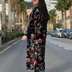 Puede incluir: Abrigo largo negro con estampado floral con detalles rojos, rosas y verdes. El abrigo se lleva con zapatillas deportivas blancas y negras. La persona lleva gafas de sol y est&aacute; de pie en una calle bordeada de palmeras.