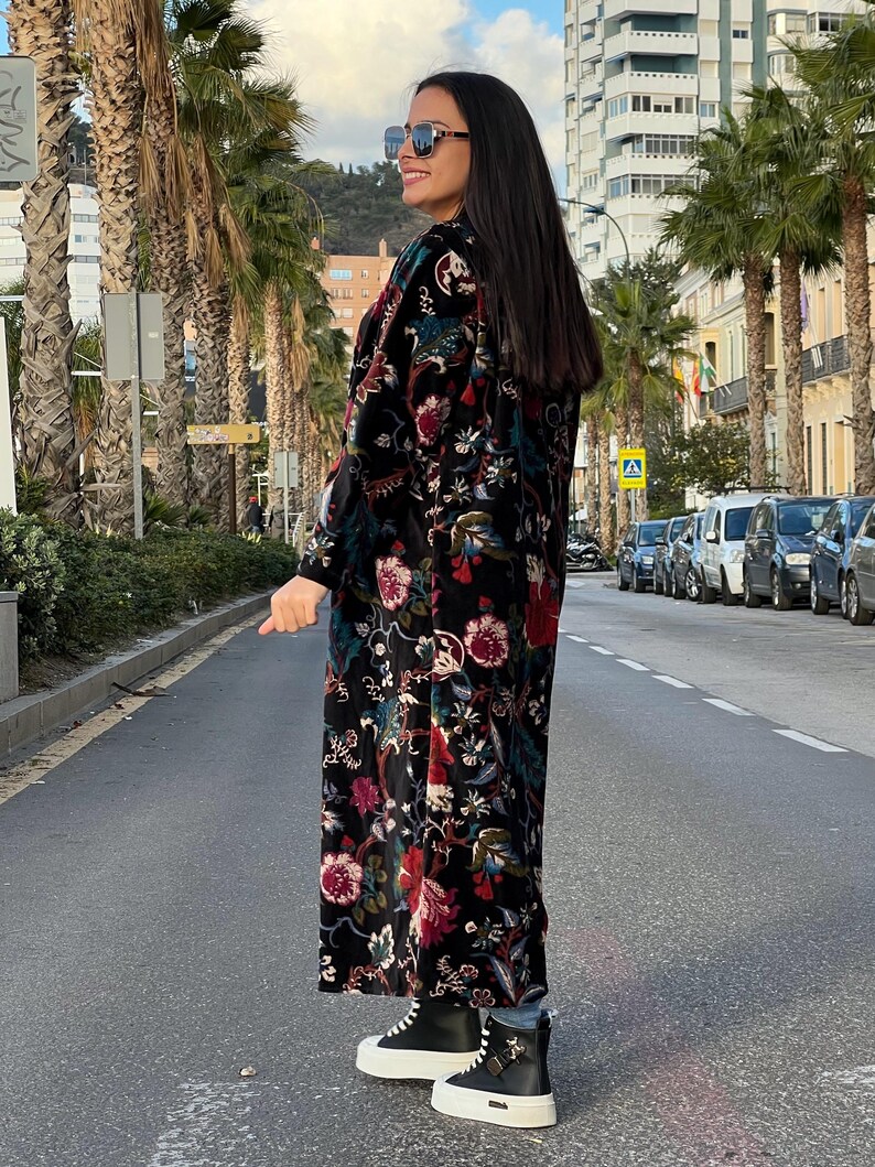 Puede incluir: Abrigo largo negro con estampado floral con detalles rojos, rosas y verdes. El abrigo se lleva con zapatillas deportivas blancas y negras. La persona lleva gafas de sol y est&aacute; de pie en una calle bordeada de palmeras.