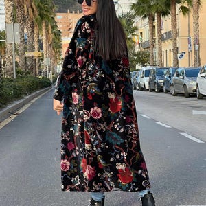 Puede incluir: Abrigo largo negro con estampado floral en rojo, rosa y verde. Se lleva sobre vaqueros azules y zapatillas de plataforma blancas y negras. El abrigo tiene una textura de terciopelo.
