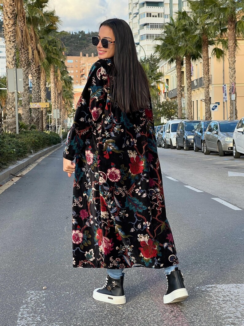 Puede incluir: Abrigo largo negro con estampado floral en rojo, rosa y verde. Se lleva sobre vaqueros azules y zapatillas de plataforma blancas y negras. El abrigo tiene una textura de terciopelo.