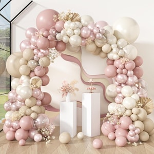 May include: A decorative balloon arch in shades of pink, cream, and beige, with clear bubble accents and dried floral embellishments. The arch is set against a backdrop with layered paper cutouts and two white pedestals.