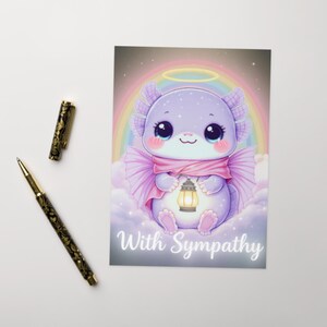 May include: A greeting card featuring a cartoon axolotl with a halo, holding a lantern, and wearing a pink scarf. The card has a rainbow background and the words "With Sympathy". A gold and black pen and pen cap are on the left.