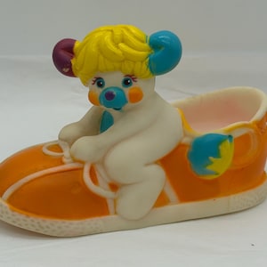 以下が含まれることがあります： オレンジ色の靴に乗った漫画のキャラクターの気まぐれな置物。キャラクターは黄色の髪、青と紫のアクセント、白い体を持っています。靴には白い靴紐とトリムが付いています。