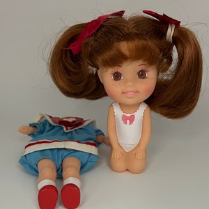 May include: A doll with brown hair in pigtails wearing a blue and white sailor dress and a white swimsuit. The doll has brown eyes and freckles.
