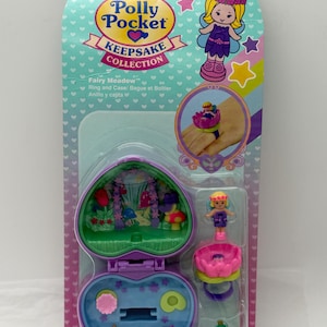 May include: A Polly Pocket Keepsake Collection Fairy Meadow ring and case set. The packaging is teal and pink, featuring a fairy illustration and a heart-shaped compact. The compact opens to reveal a miniature fairy scene with a pond and a miniature fairy figure.
