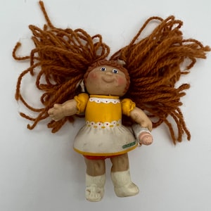 Op de afbeelding: Een vintage Cabbage Patch Kid pop met garenhaar, die een geel en wit jurkje draagt en een flesje vasthoudt.