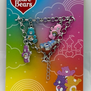 Könnte beinhalten: Ein silberfarbenes Charm-Armband mit Care Bear-Anhängern. Das Armband hat fünf farbenfrohe Care Bear-Anhänger, darunter ein blauer, rosa, lila und zwei weitere Bären. Der Hintergrund ist ein Regenbogenverlauf mit Care Bears-Branding.