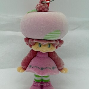 May include: A vintage Strawberry Shortcake doll figurine. The doll has a pink head shaped like a strawberry, with green stripes and a red berry on top. She wears a pink dress, red sleeves, and green and white striped socks. The doll is a collectible item.