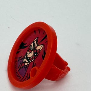 May include: A bright red plastic ring featuring a circular face with a graphic illustration of a character. The design includes a small circular cutout and the text 1987 TELEPIX printed along the inner edge of the red frame surrounding the image.