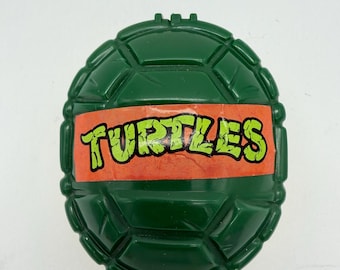 May include: A green, turtle-shell-shaped toy with a textured surface. An orange band across the center displays the word "TURTLES" in green, stylized lettering. The toy is likely a collectible or part of a larger set.