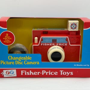May include: A vintage Fisher-Price toy camera in its original box. The red camera has a white lens and a flash cube. The box is red, blue, and yellow, with the text "Changeable Picture Disc Camera" and "Fisher-Price Toys".