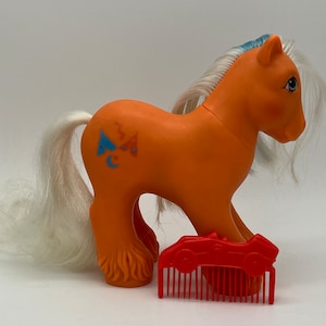 Puede incluir: Pony My Little Pony naranja con una melena y cola blancas. El pony tiene un símbolo azul, naranja y blanco en su flanco. El pony sostiene un coche de carreras rojo de juguete y un peine rojo.