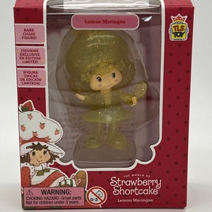 May include: A yellow and green plastic Strawberry Shortcake doll with a lemon meringue theme. The doll is holding a small mirror. The packaging is red with white polka dots and features the text "The World of Strawberry Shortcake" and "Lemon Meringue".