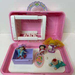 Pode incluir: Uma mala rosa Cabbage Patch Kids, aberta para revelar um interior branco com várias figuras. A mala tem uma alça e um topo decorativo. No interior existem várias bonecas e um pequeno tapete.
