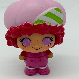 May include: A small, colorful figurine of a character with a pink strawberry-shaped hat and red curly hair. The character has large purple eyes, freckles, and a smiling expression. The outfit is a magenta dress with heart details.