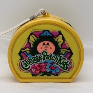 May include: A yellow Cabbage Patch Kids carrying case with a white strap. The front features a cartoon image of a Cabbage Patch Kid with the words "Cabbage Patch Kids" in a green, pink, and blue design. The case is from 1983.