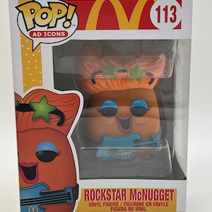 May include: A Funko Pop! Ad Icons vinyl figure of a Rockstar McNugget. The McNugget is wearing a blue shirt, a green star headband, and is holding a blue guitar. The figure is in a clear plastic box with a white background. The box has the McDonald's logo and the words "Rockstar McNugget" on it.