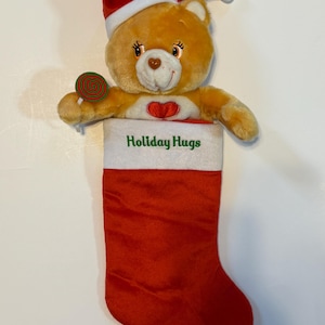 Könnte beinhalten: Ein beiger Teddybär mit roter Weihnachtsmütze und Lutscher, der aus einem roten Weihnachtsstrumpf kommt. Der Strumpf hat einen weißen Bund mit den grünen Worten "Holiday Hugs".